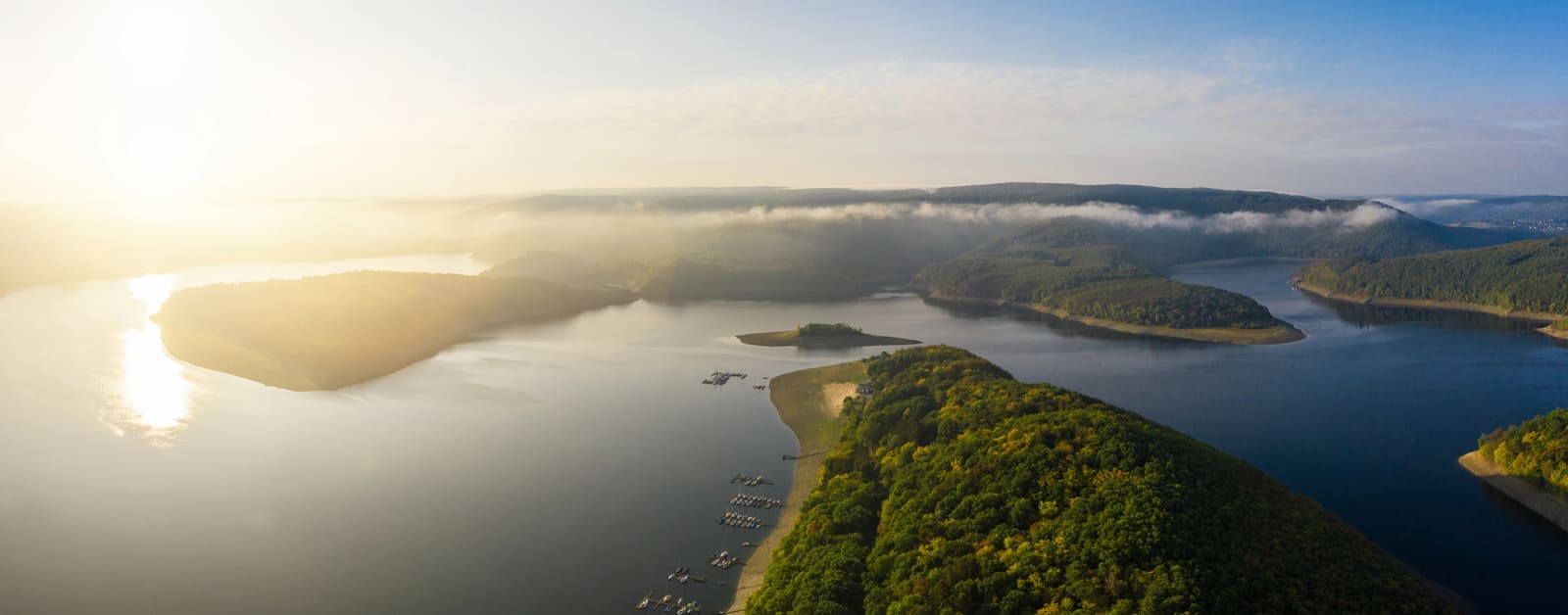 Luftpanorama des Rursees bei Sonnenaufgang mit bewaldeten Halbinseln, kleinen Marinas und Nebelbändern über den Eifelhöhen