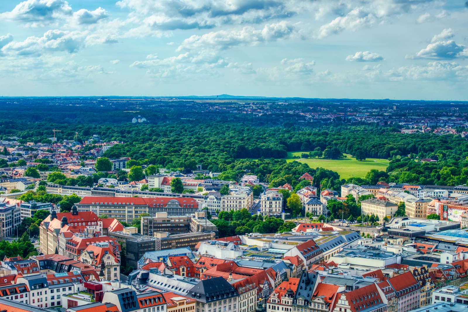 Luftblick über Leipzigs dicht bebaute Innenstadt mit roten Dächern und dem grünen Clara-Zetkin-Park am Horizont