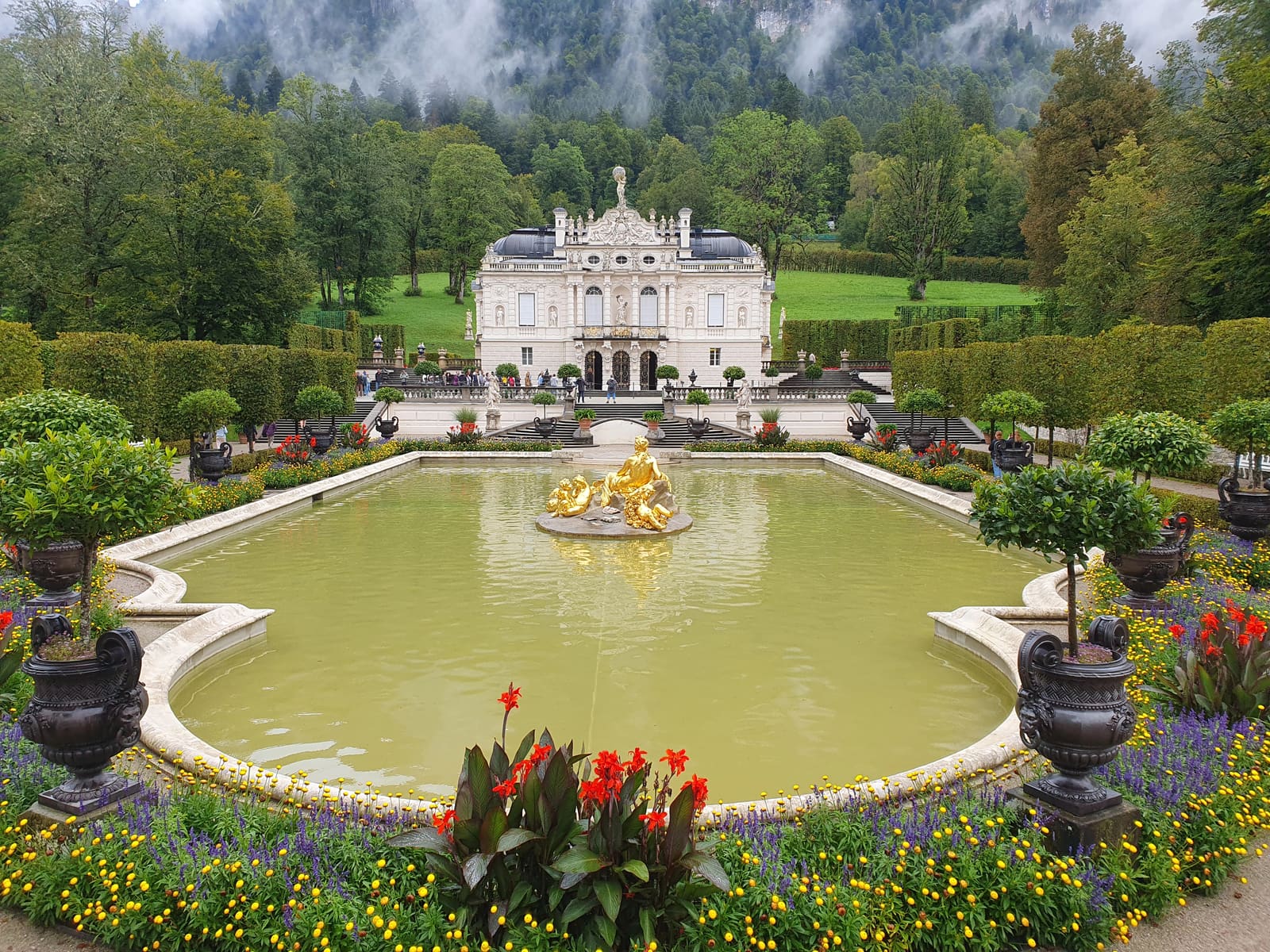 Schloss Linderhof hinter dem Wasserparterre mit vergoldeter Figur, bunten Blumenbeeten und bewaldeten Berghängen