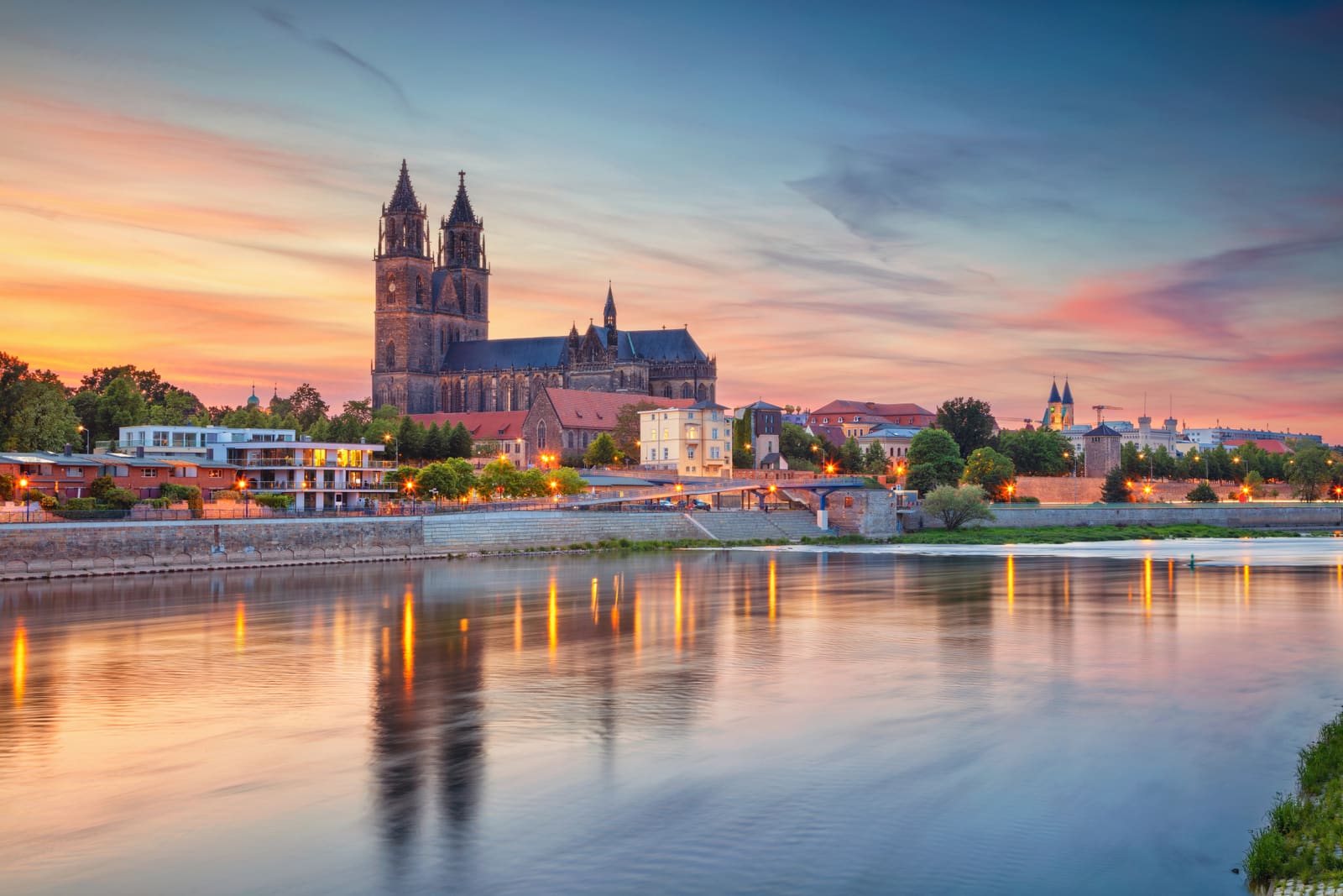 Magdeburger Dom und beleuchtete Elbuferbebauung vor rosa-orangefarbenem Abendhimmel mit Spiegelungen im ruhigen Fluss