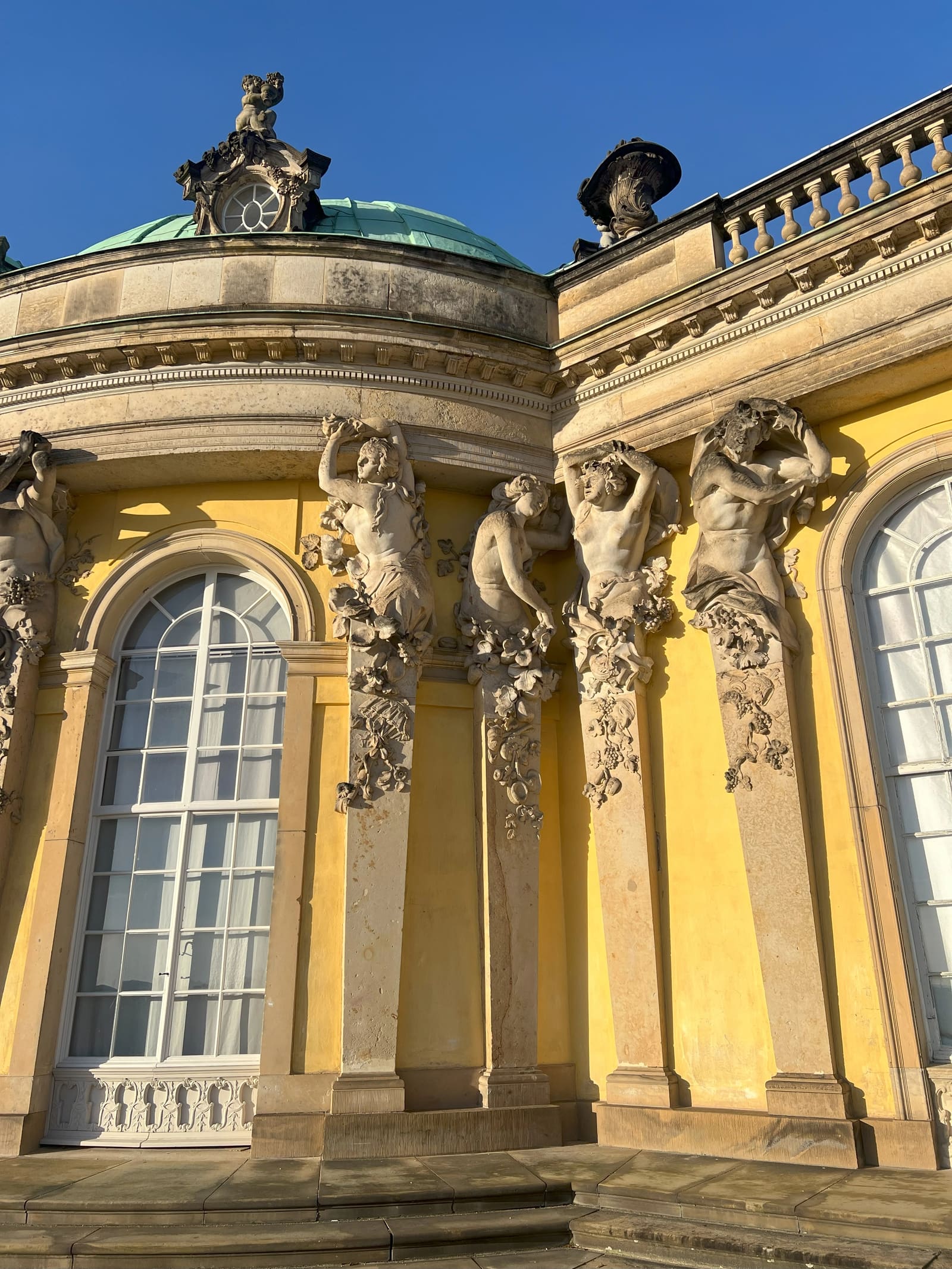 Gelbe Fassade von Schloss Sanssouci mit hohen Rundbogenfenstern, steinernen Karyatiden und kupfergrünem Kuppeldach