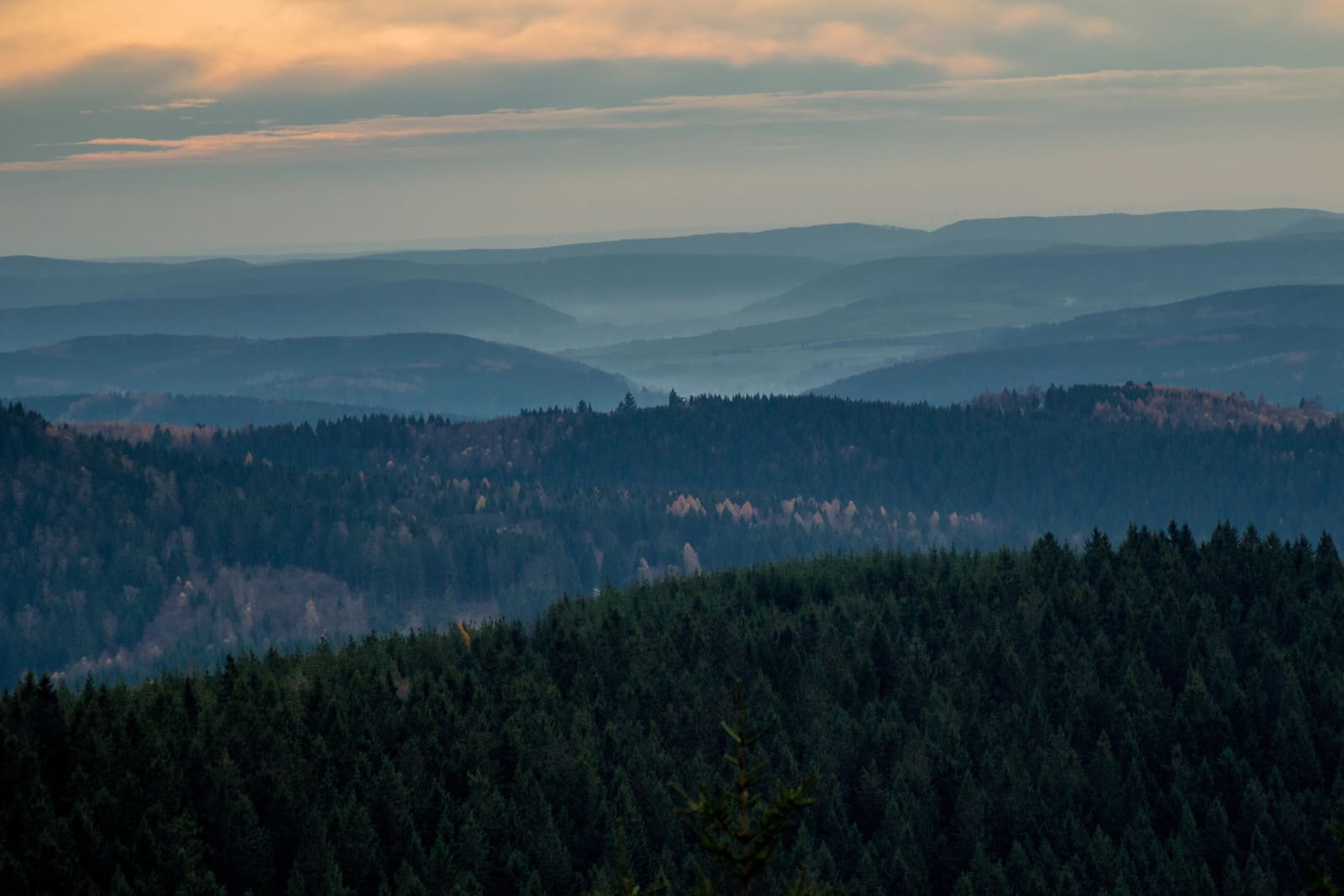 Gestaffelte bewaldete Bergrücken des Thüringer Walds am Rennsteig im Dunst unter orange beleuchteten Abendwolken
