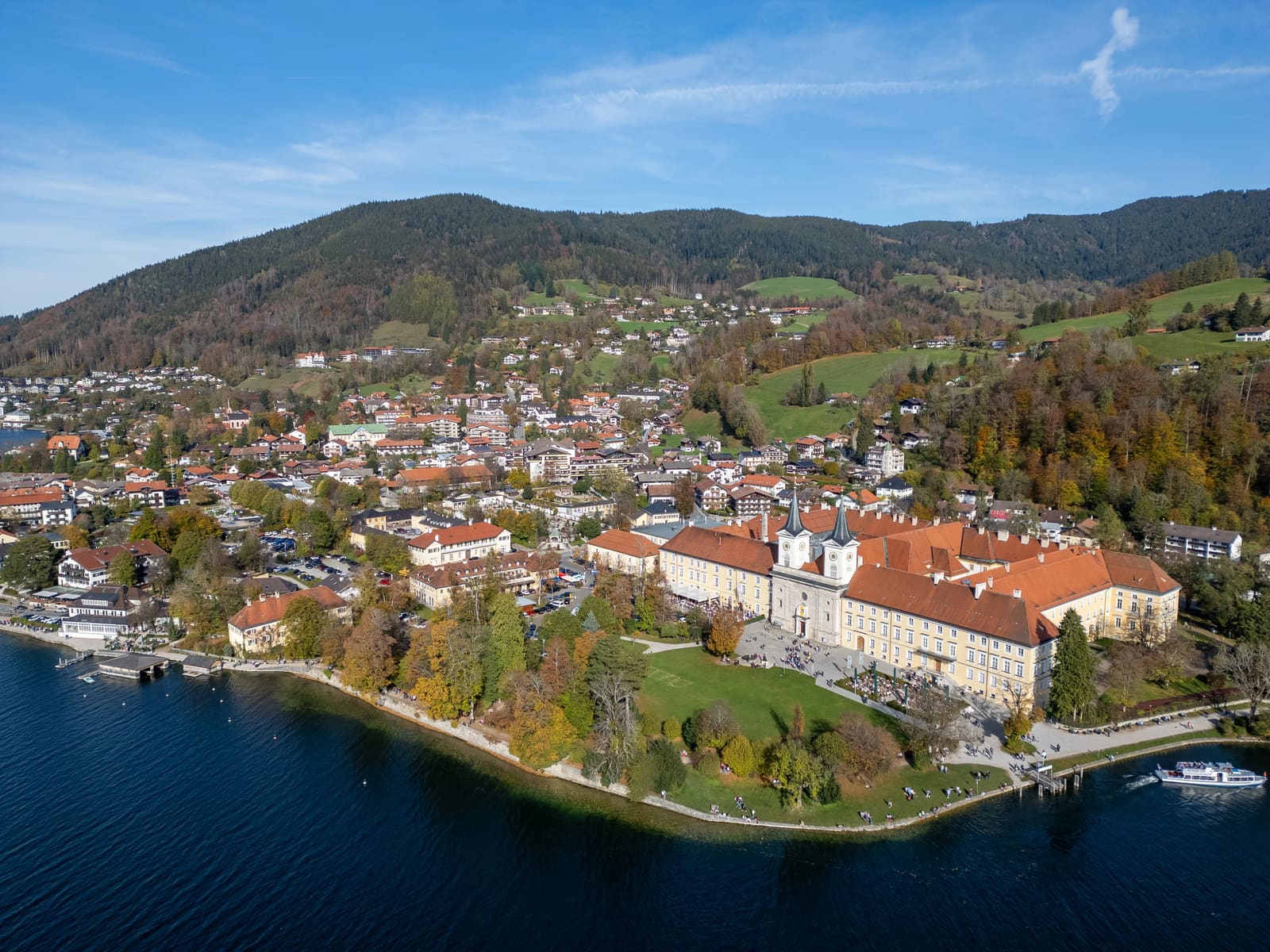 Luftbild des ehemaligen Klosters Tegernsee mit Doppeltürmen, Uferpromenade, Stadt und bewaldeten Hängen