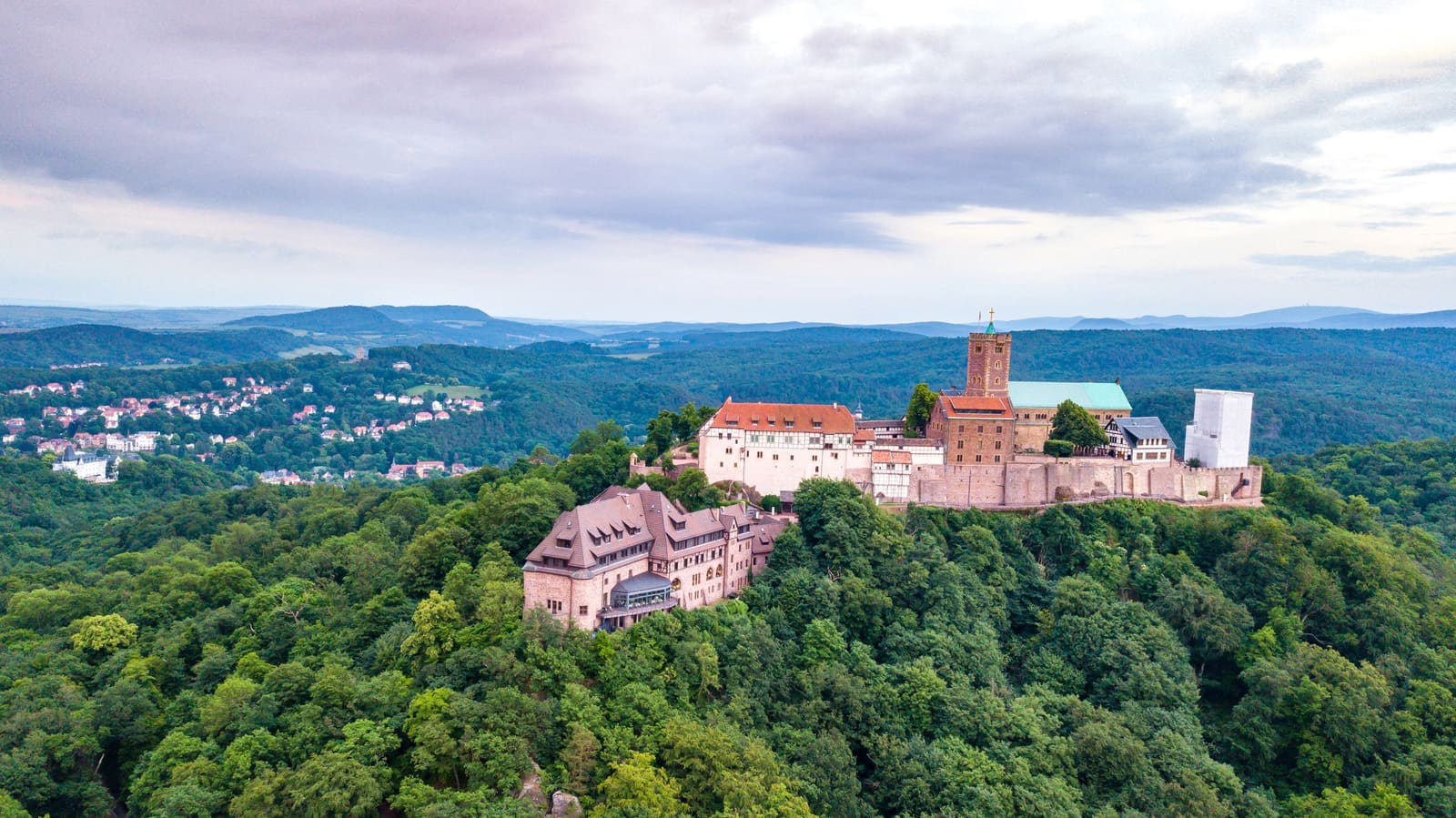 Luftbild der Wartburg auf einem dicht bewaldeten Höhenzug mit Eisenach und den Hügeln des Thüringer Walds im Hintergrund