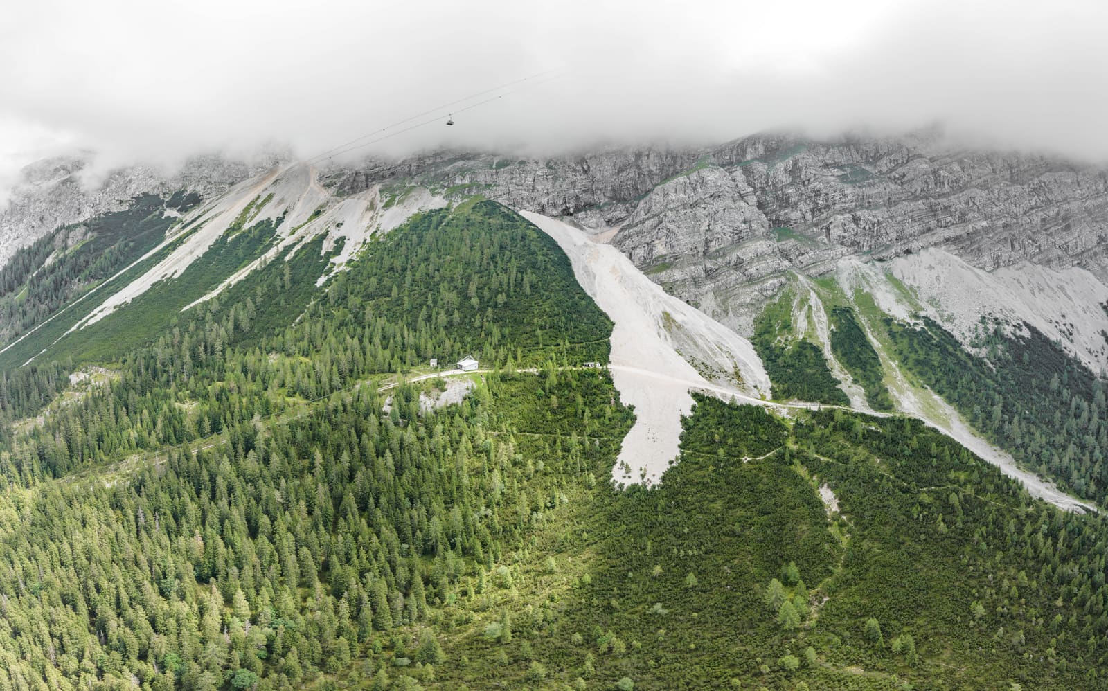 Luftbild der bewaldeten Riffelriss-Route unter dem wolkenverhangenen Zugspitzmassiv mit Bahntrasse und kleinen Stationsgebäuden
